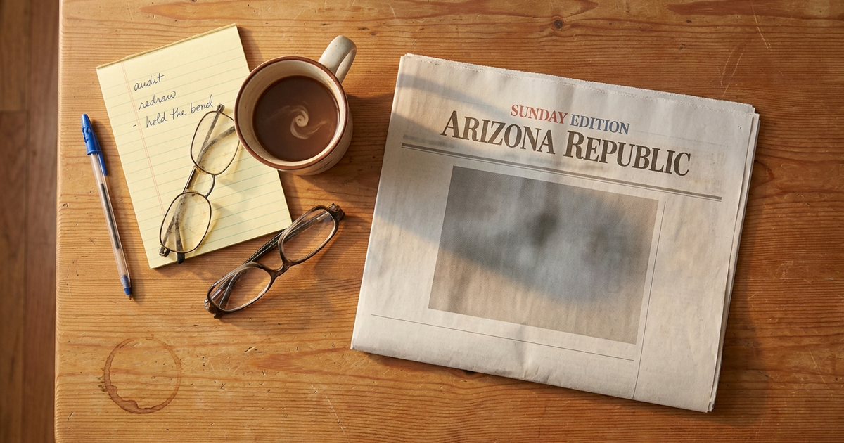 Sunday edition of the Arizona Republic folded on a pine kitchen table next to a coffee mug, reading glasses, and a yellow legal pad.